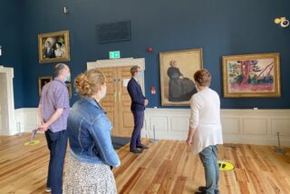 A group of four people wearing masks stand in the Captain's Room with wooden floors and blue walls, observing paintings. The atmosphere is quiet and contemplative.