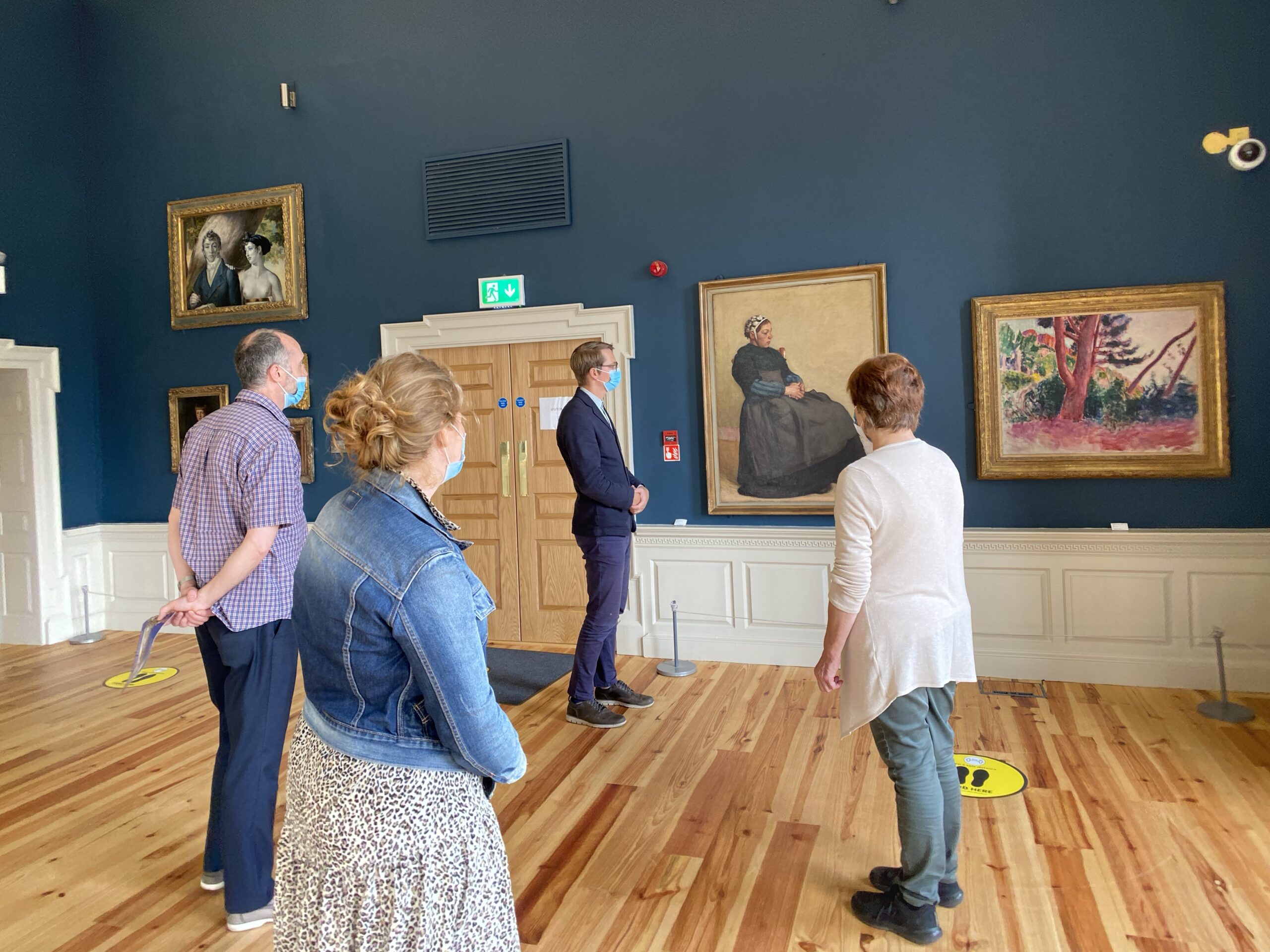 A group of four people wearing masks stand in the Captain's Room with wooden floors and blue walls, observing paintings. The atmosphere is quiet and contemplative.