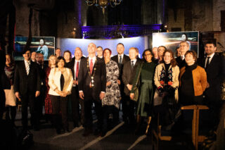 A diverse group of people, dressed formally, smiling and posing together in a dimly lit room with a warm atmosphere. Screens in the background display a woman's portrait.