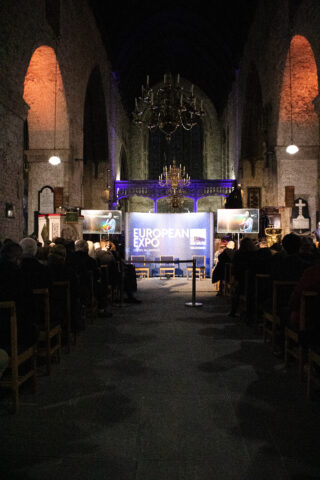 A dimly lit church with arched ceilings and illuminated stone walls features a central screen displaying a presentation, creating a solemn and focused atmosphere.
