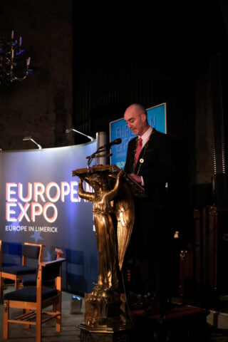 A person stands at a podium adorned with a golden eagle, speaking at the European Expo in Limerick. The backdrop features an illuminated sign.