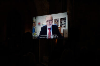 A man in a suit and red tie speaks on a video screen in a dimly lit room. The subtitle reads: 