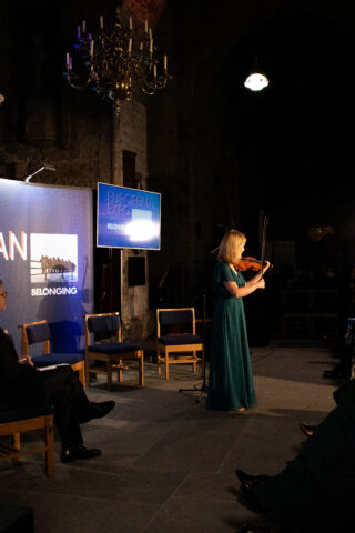 A woman in a green dress plays the violin on stage under dim lighting. A 