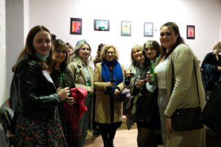 A group of people smiling at a social gathering, holding drinks. Artwork is visible on the wall behind them. Warm, friendly atmosphere.