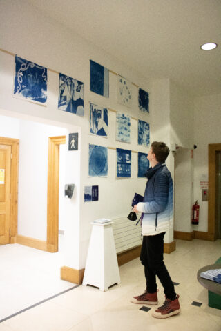 A person in a gradient blue and white jacket observes cyanotype prints on a white wall in a well-lit gallery. The mood is calm and contemplative.