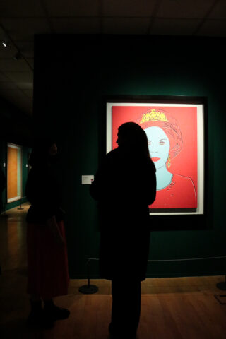 Silhouetted figures stand in a dimly lit gallery, viewing a vibrant framed portrait featuring a stylized woman with a crown against a red background.