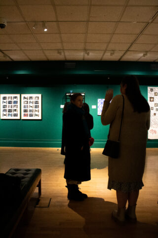 Three people conversing in a dimly lit art gallery with green walls. Framed artworks are visible in the background, creating a contemplative mood.