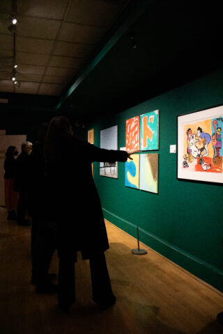 Visitors in a dimly lit art gallery observe vibrant, abstract paintings on a teal wall. One person points, creating an atmosphere of engagement.