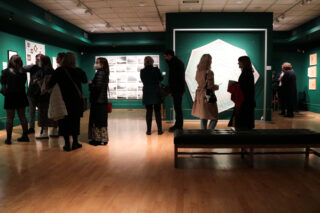 A group of people in a dimly lit art gallery, admiring artwork on green walls. They are wearing coats, some with masks, conveying an atmosphere of quiet focus.