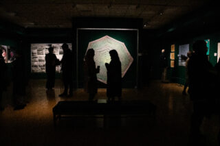 Dimly lit art gallery with silhouettes of people viewing displays. Central focus on a large hexagonal art piece, creating a contemplative atmosphere.