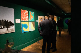 People in an art gallery with teal walls, viewing colourful abstract paintings. The scene is dimly lit, and a glass of white wine is on a table.