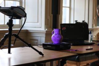 Person in a mask operates a laptop and 3D scanner, capturing a purple clay jug on a turntable. Soft natural light filters through a window.