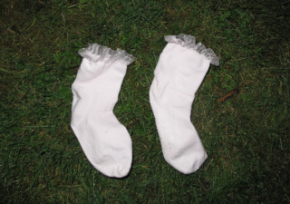 Two white socks with ruffled lace cuffs are placed on lush green grass. The image conveys a whimsical, playful tone against a natural backdrop.