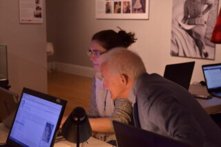 Two people, a woman and an older man, focus intently on laptops in a dimly lit room. They're collaborating, creating an atmosphere of concentration.
