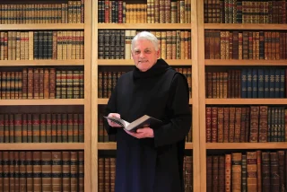 Patrick Hederman in a black robe stands holding a book, surrounded by tall wooden shelves filled with numerous antique books.