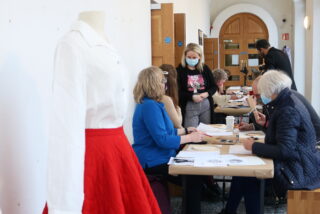 Mannequin in white blouse and red skirt in the foreground. People seated at a table in the background, engaging in a creative workshop. Bright setting.