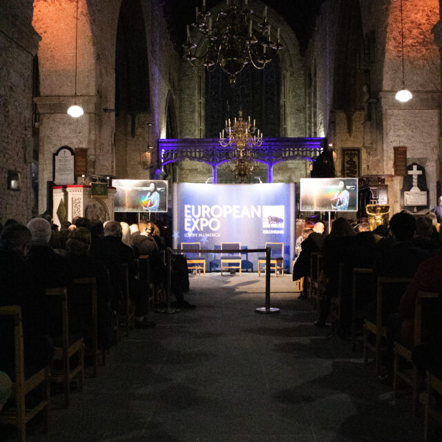 A dimly lit church interior hosts the European Expo event. Attendees sit on wooden chairs, facing a blue stage with displays and chandeliers overhead, creating a solemn atmosphere.