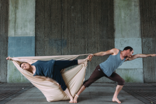 Two dancers perform under a bridge. One is wrapped in fabric, held by the other in an extended pose. The scene conveys balance and tension.