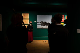 A person in a mask and school uniform presents in a dimly lit gallery. Behind them, art pieces hang on a green wall, observed by silhouetted figures.