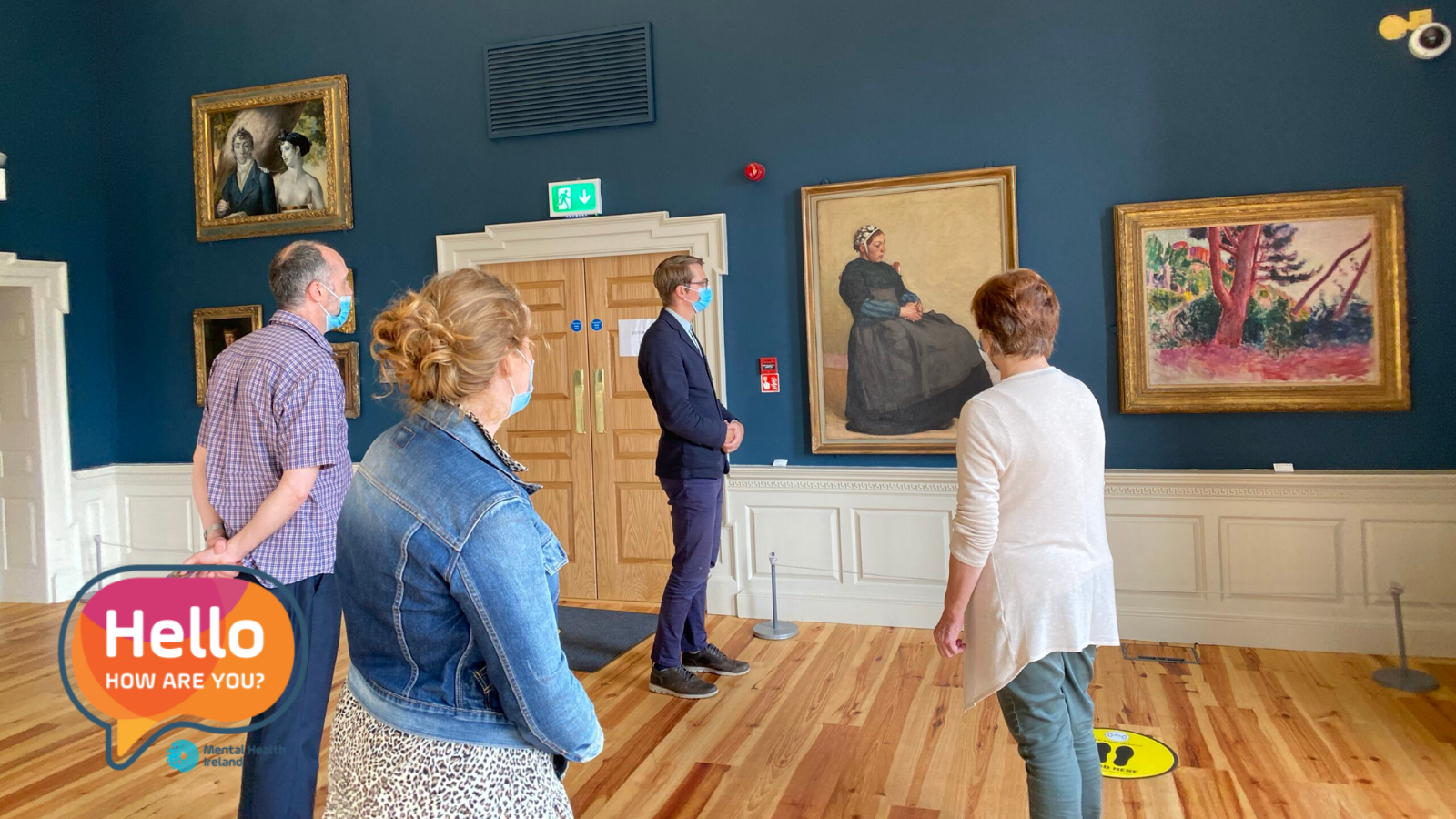 A small group of people, wearing masks, stands in a gallery observing paintings on a blue wall. A 