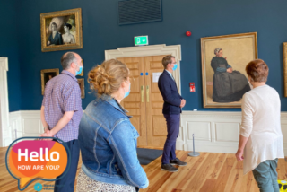 A small group of people, wearing masks, stands in a gallery observing paintings on a blue wall. A 