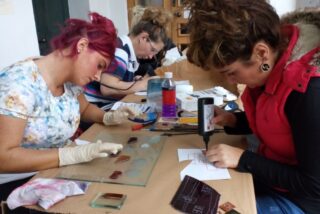 Three women focused on creating stained glass art at a table. One is drawing on paper, another handling small glass pieces. Tools and supplies are scattered.