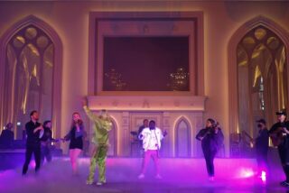 A band performs energetically in a grand hall with gothic-style windows. The scene is illuminated with purple and gold lights, creating a lively atmosphere.