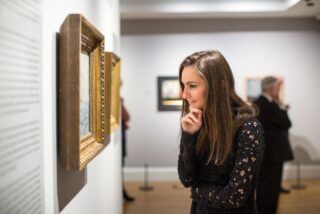 A woman in a black lace top thoughtfully observes a framed painting in a museum, conveying curiosity. Blurred background shows other visitors.