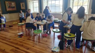 A classroom with students in uniform working on art projects. They sit at round tables with colourful stools, surrounded by natural light and wooden floors.