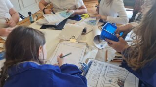 Group of children in blue smocks engaged in artwork at a table. They use various art supplies like brushes and colouring sheets, creating a creative, lively atmosphere.
