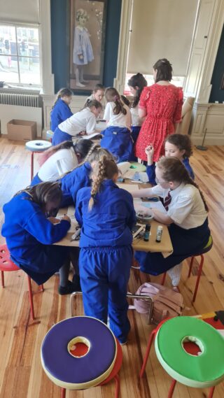 A group of children in blue uniforms huddle around a table, focused on an art project. An adult in red assists. The room is bright and airy.