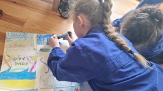 Two children in blue shirts are lying on wooden floorboards, colouring a bright, detailed drawing with trees, a river, and a castle. Their expressions are focused.