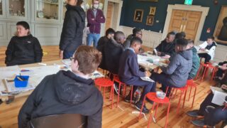 A group of people seated around tables in a room engaged in activities. Some stand; others sit on red stools. The atmosphere is focused and collaborative.