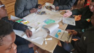 A group of people gather around a table filled with papers, drawings, and art supplies, engaged in creative activities. The mood is collaborative and focused.
