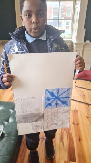 A child stands indoors, holding a poster with a detailed drawing. The room is bright with natural light from a window, creating a focused and proud atmosphere.