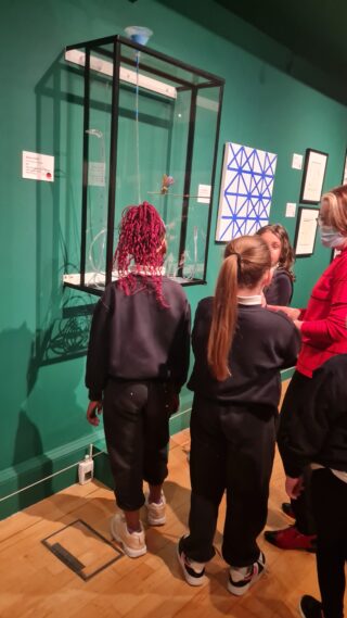 Children in a museum gallery examine art displayed in a glass case. The room has green walls with framed artworks, creating an atmosphere of curiosity and learning.