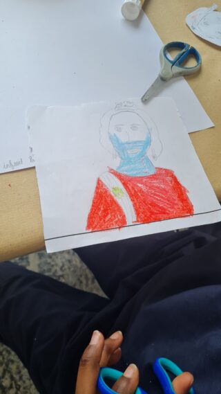 A child's hand holds blue scissors near a colourful drawing of a person in a red outfit. Papers and a tape dispenser are on the table.