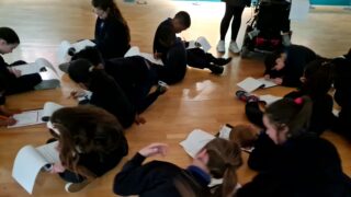 Children sit on a wooden floor in a group, focused on reading and writing in their notebooks, creating a calm and studious atmosphere.