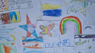 Children's colourful drawings on white paper include a butterfly, rainbow, star, and scribbles. The mood is cheerful and playful.