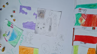 Children's colourful drawings scattered on a table, featuring flags, abstract shapes, and doodles. The scene conveys a creative and playful atmosphere.