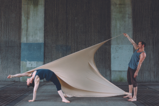Cikada Circus Performers, Two people perform a unique dance. One bends backward, draped in beige fabric, while the other holds the fabric taut, creating dynamic tension.