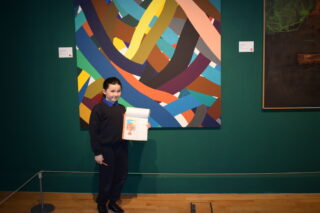 A child stands in a gallery holding a drawing, smiling. Behind, a colourful abstract painting contrasts with green walls, creating a lively, creative atmosphere.