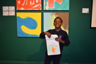 A young boy smiles proudly, holding a colourful drawing in front of abstract art pieces on a gallery wall. The scene conveys joy and creativity.