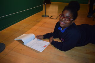 A person lies on a wooden floor, smiling and reading an open book, wearing glasses and a dark sweater. The background features a green wall, creating a cosy, relaxed atmosphere.