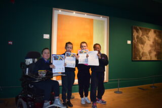 Four young people stand proudly holding awards in front of a vibrant orange wall. A person in a wheelchair is smiling beside them. The setting is warm and celebratory.