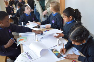 Children in school uniforms sit at a table engaged in a group art project. Papers, markers, and glue are scattered around, conveying a creative atmosphere.