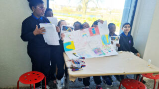A group of children in uniforms proudly displays a colourful poster with drawings and photos. They are gathered around a table, smiling and showing teamwork.