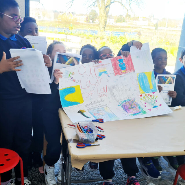 A group of smiling children in school uniforms present colourful posters at a table. Bright sunlight streams through a window, creating a joyful atmosphere.