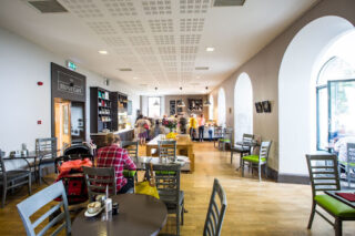 Spacious Hunt café with arched windows and wooden floors. Patrons sit at grey tables, enjoying a cosy atmosphere. Bright, inviting, and bustling.