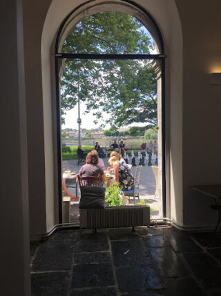 View through an arched window of two people seated at a sunlit outdoor café table, under a tree, with a scenic background and people walking nearby.